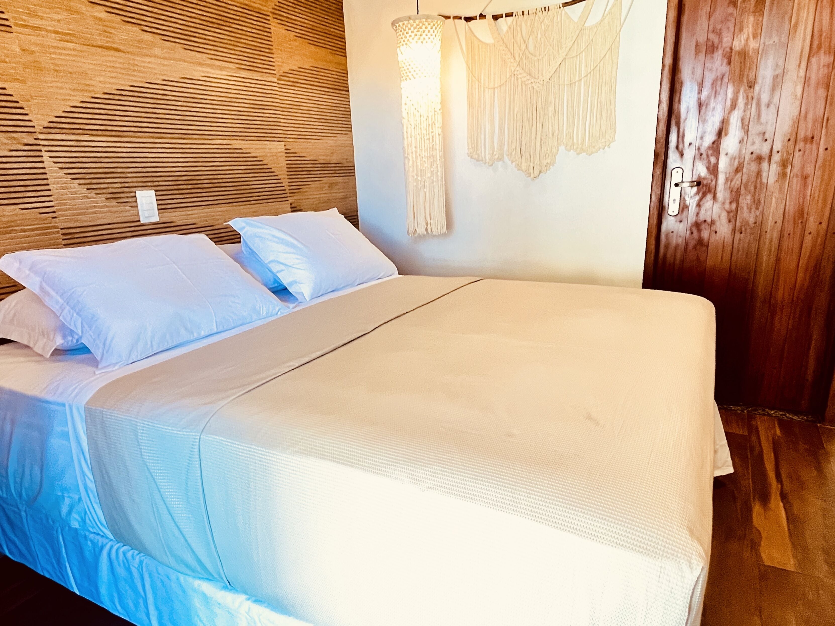 Una cama con ropa blanca en una habitación con paneles de madera y decoración de macramé en la pared.
