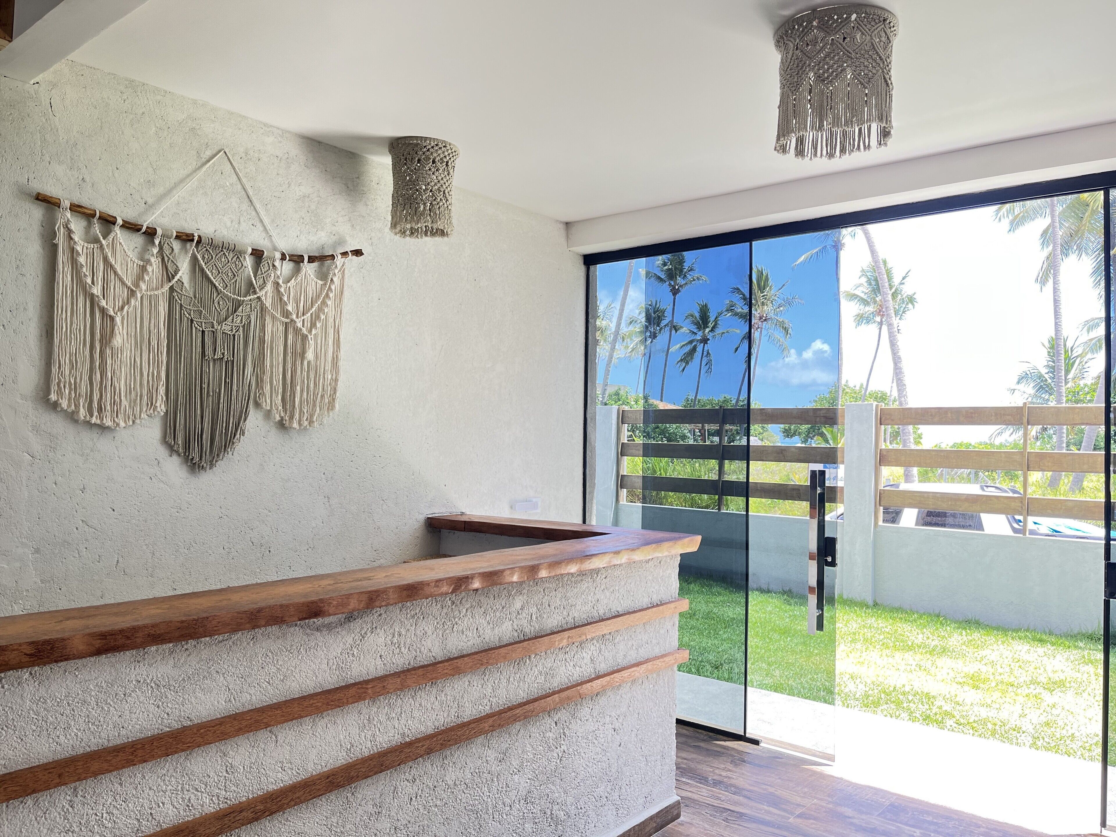 Un área de recepción moderna con un mostrador de concreto, decoración de pared con macramé y vistas a palmeras a través de grandes puertas de vidrio.