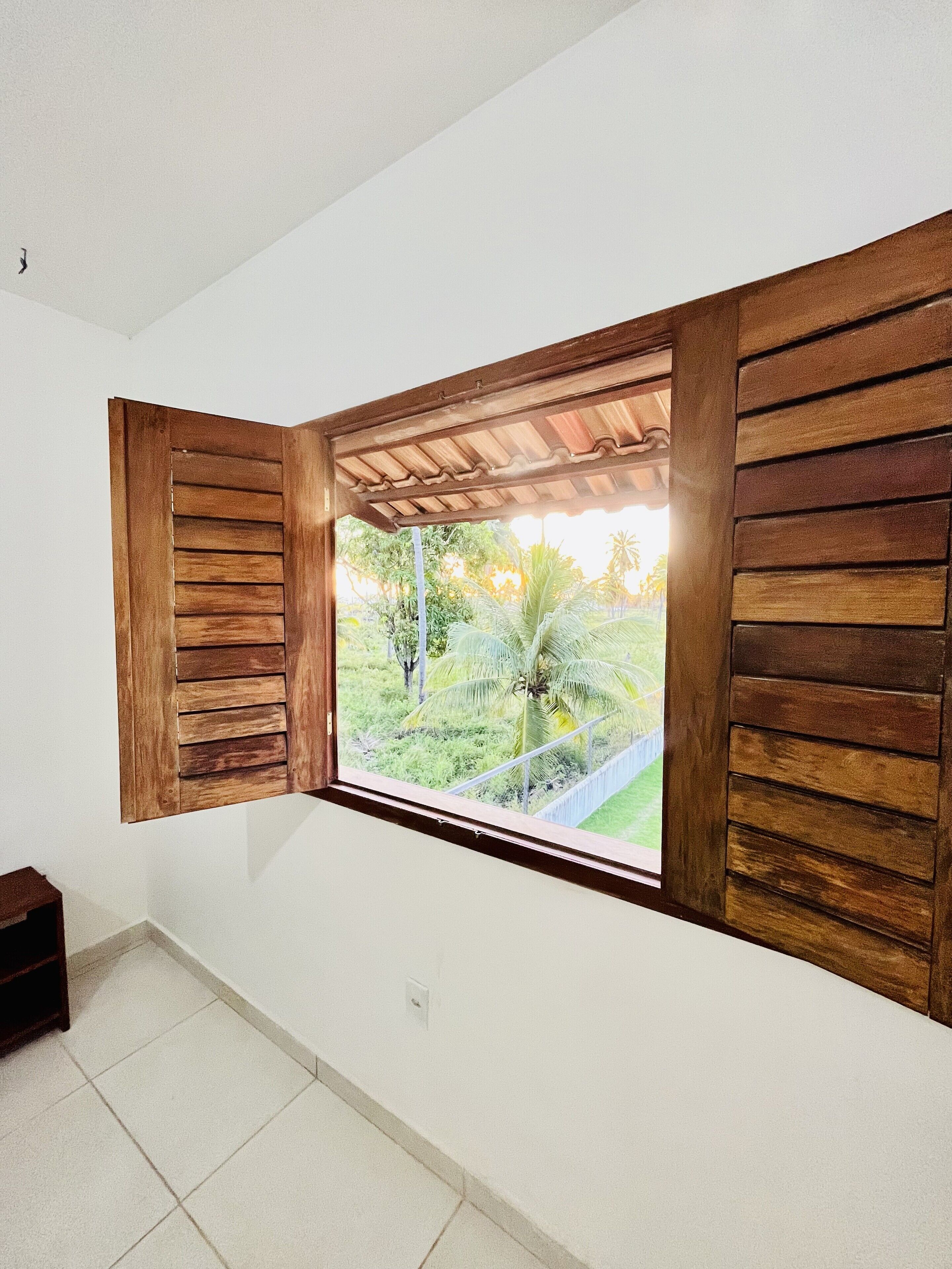 A room with a window featuring wooden shutters, a white wall, and a tiled floor.