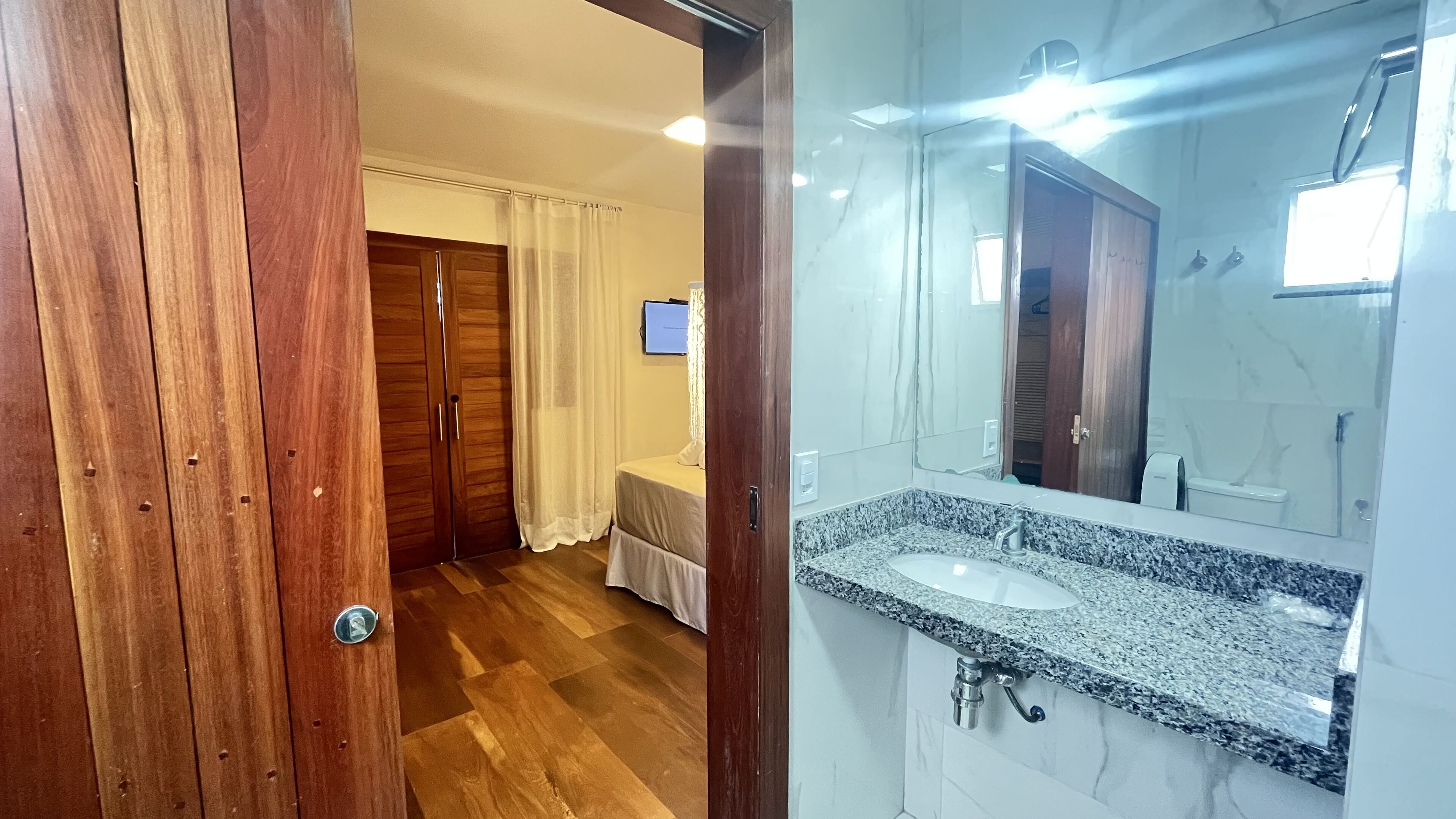 Habitación de hotel con armario de madera, cama con cubrecama blanco, y baño con encimera de granito y espejo.