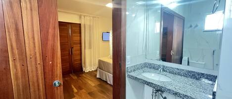 Habitación de hotel con armario de madera, cama con cubrecama blanco, y baño con encimera de granito y espejo.