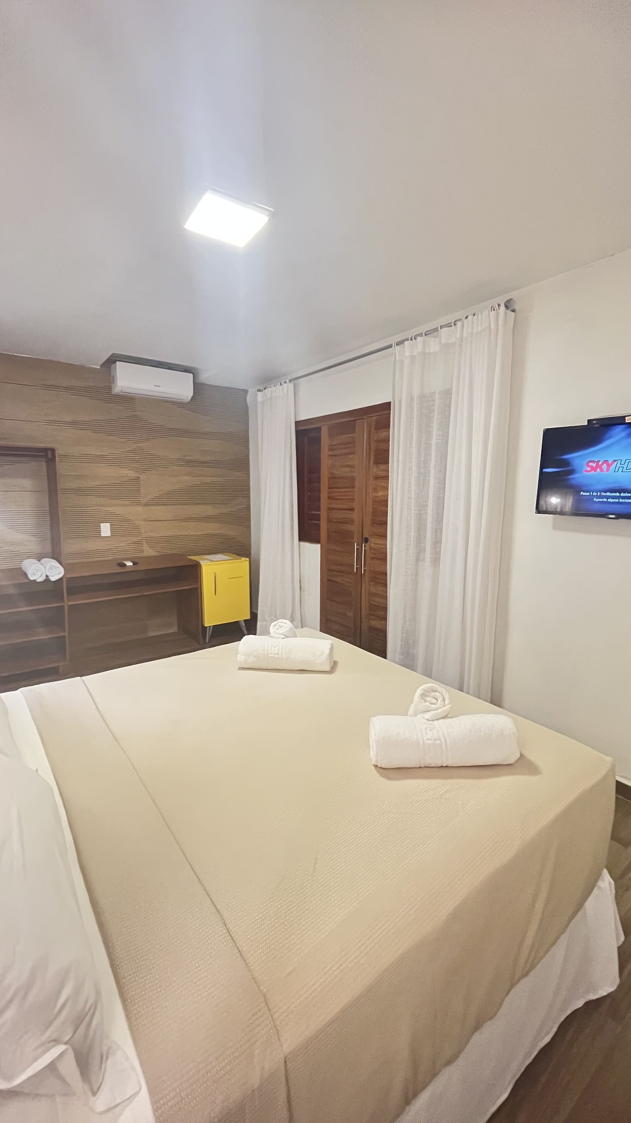 Una habitación de hotel con una cama grande, un armario de madera, un mueble amarillo y un televisor en la pared.