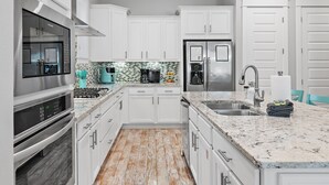 A modern kitchen with white cabinets, stainless steel appliances, a granite countertop, and a patterned backsplash. - New Golf Cart | Resort Style Pool | 3 Bikes | 30A (Panama City Beach)