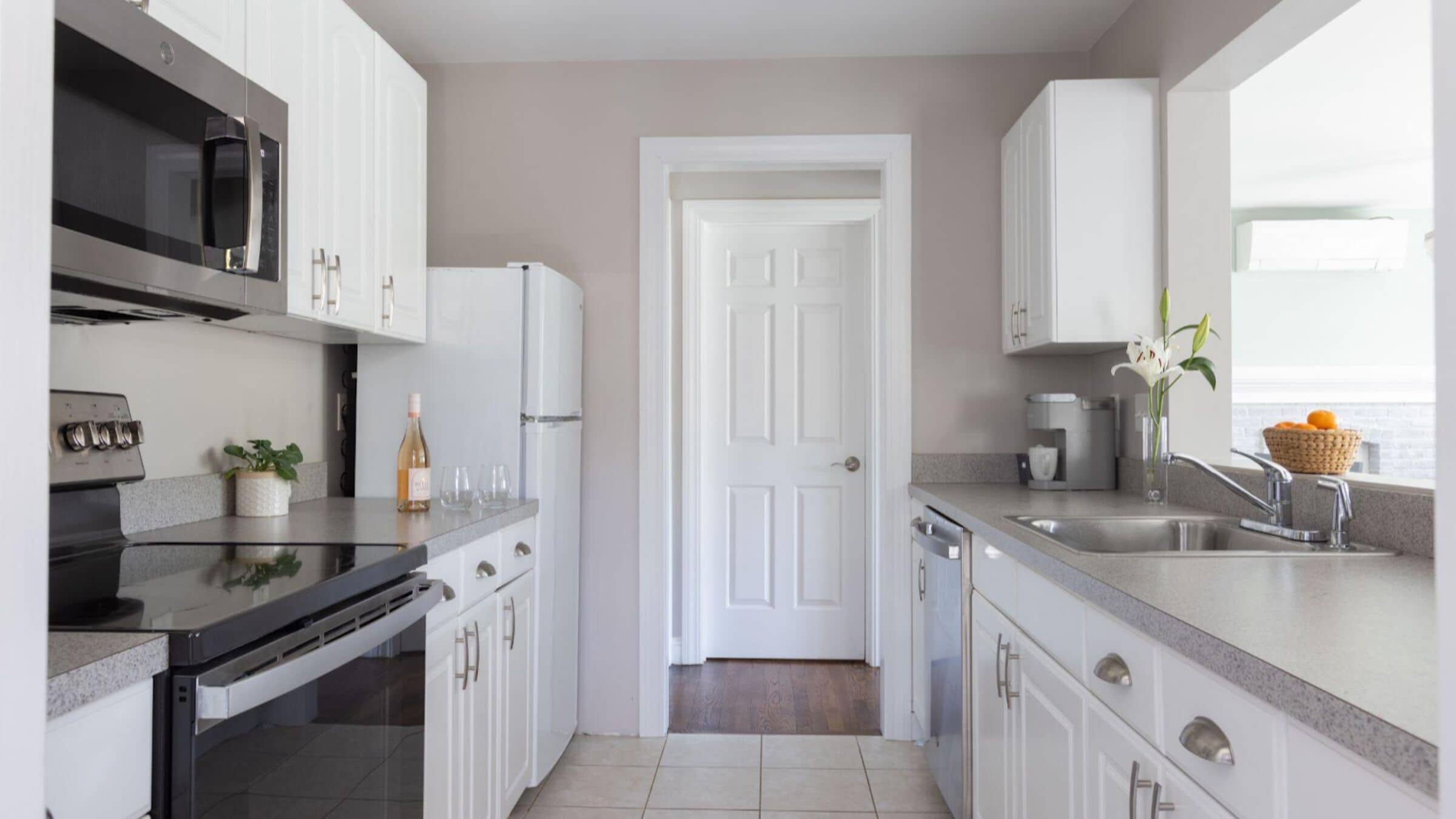 A kitchen with white cabinets, a microwave, a refrigerator, a sink, and a window with a basket of oranges.