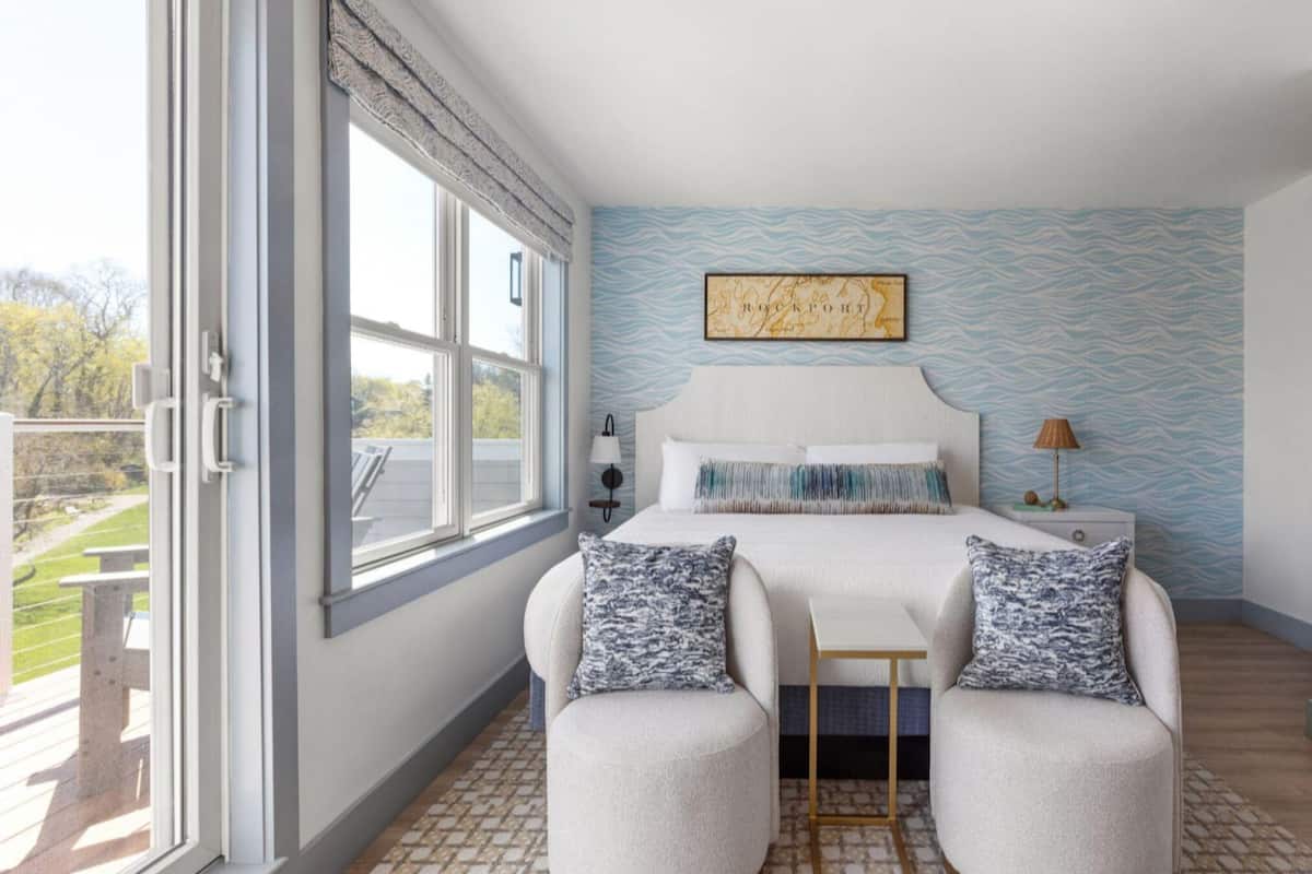 A bedroom with a bed, two chairs, a nightstand, a lamp, and a window with a view.