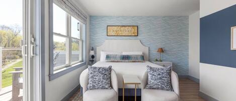 A bedroom with a bed, two chairs, a nightstand, a lamp, and a window with a view.