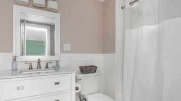 A bathroom with a white sink, mirror, and toilet, a basket on the toilet, and a shower curtain.