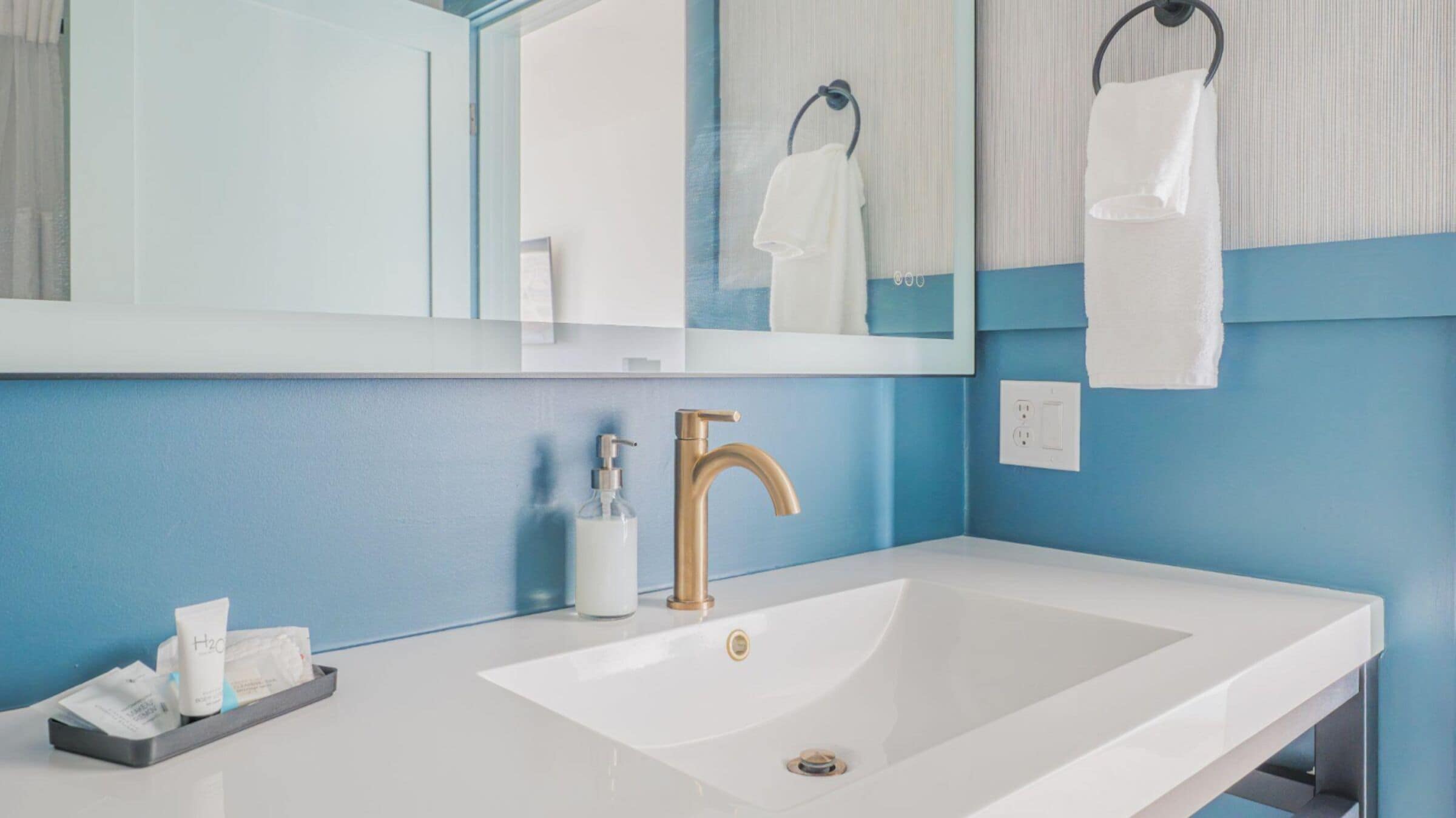 A bathroom with a white sink, a gold faucet, a mirror, and a towel rack.