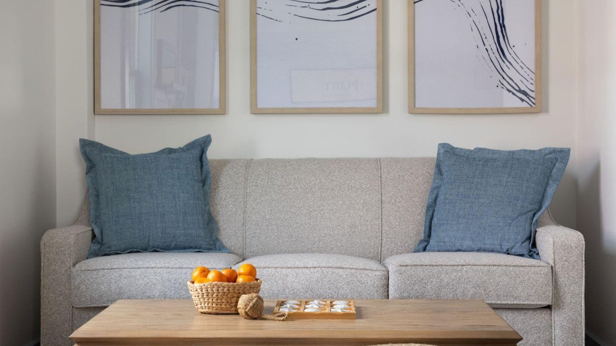 A living room with a grey sofa, two blue cushions, a wooden coffee table, a basket of oranges, and a woven basket.