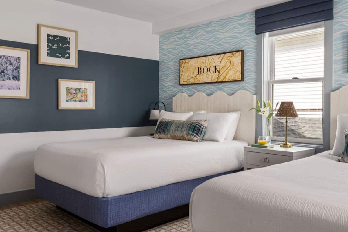 A hotel room with two beds, a window with blinds, wall art, and a nightstand with a lamp.