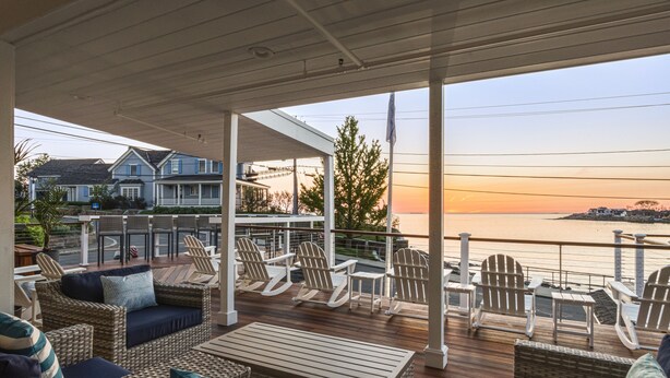 A covered outdoor deck with wicker furniture, a view of the sea, and houses in the background. - The Cove at Rockport Hotel (Rockport)