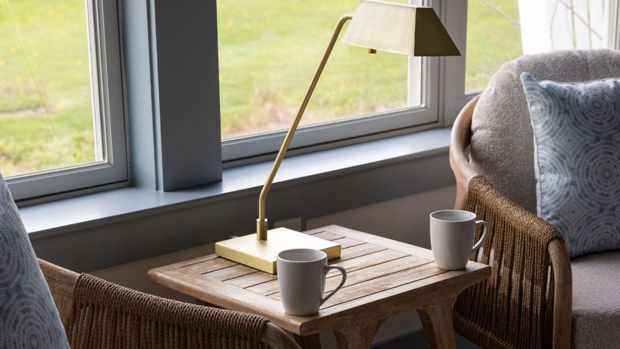 Una habitación acogedora con una mesa de madera, dos tazas blancas, una lámpara y vistas a la vegetación a través de una ventana.