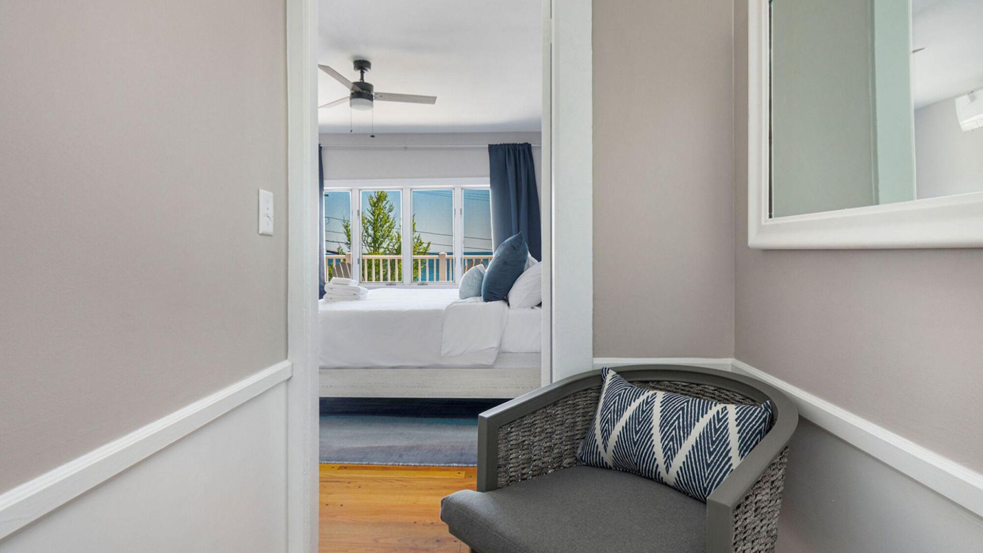 A narrow hallway leading to a balcony with a bed, a ceiling fan, and a chair with a patterned cushion.
