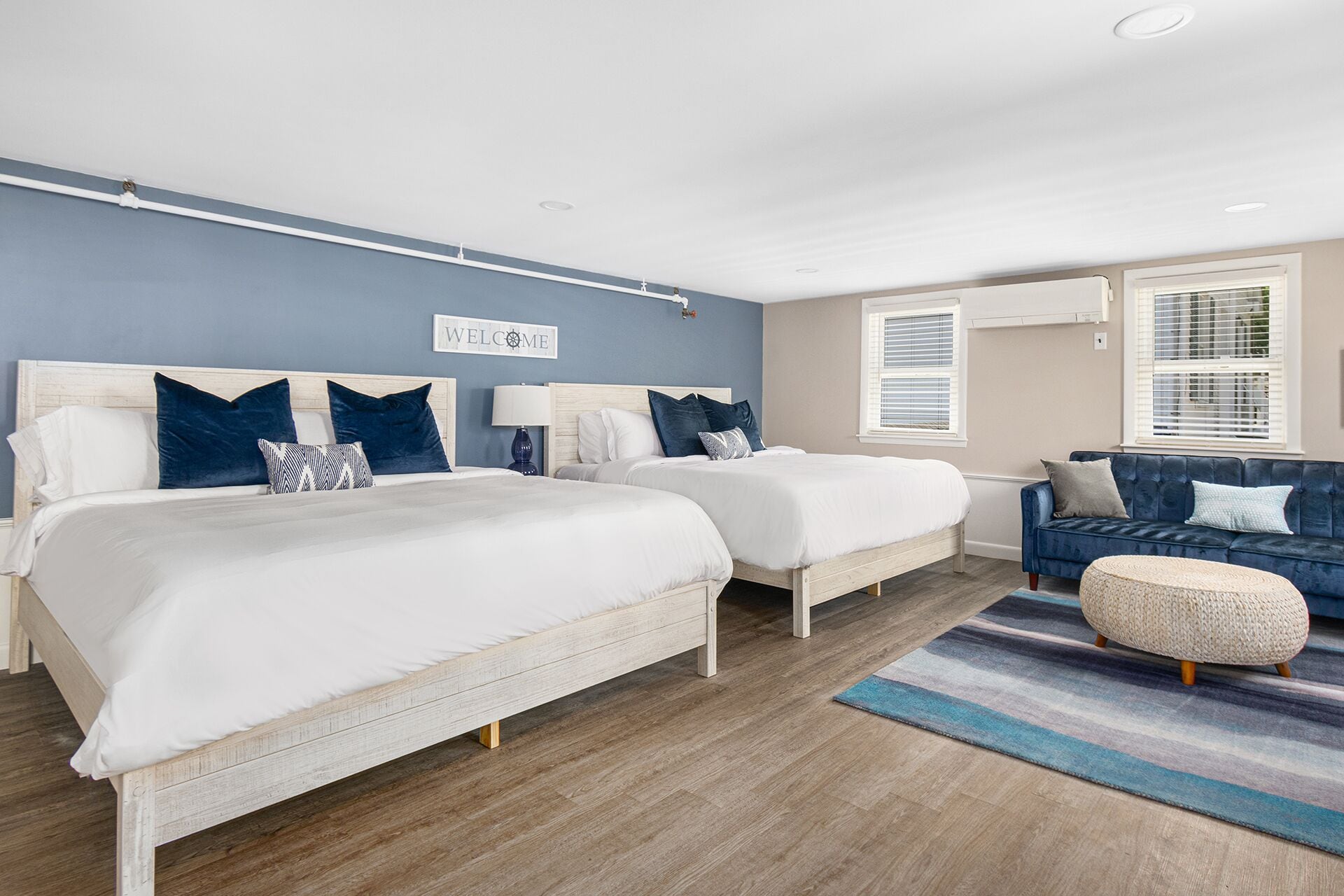A modern bedroom with two beds, a blue sofa, and a 'WELCOME' sign on the wall.