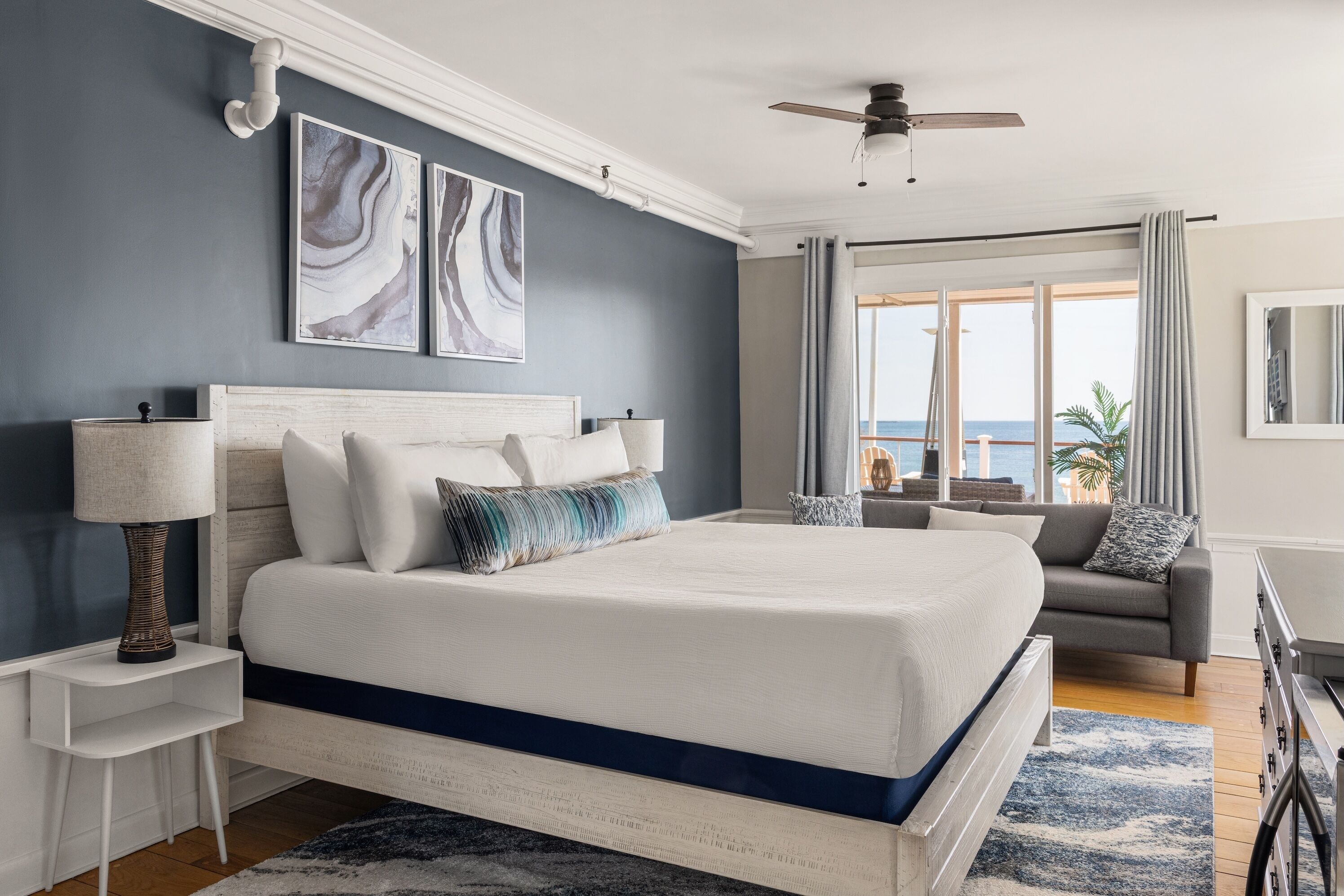Quarto com uma cama grande, mesa de cabeceira, luminária, ventilador de teto, janela com cortinas e obras de arte na parede.