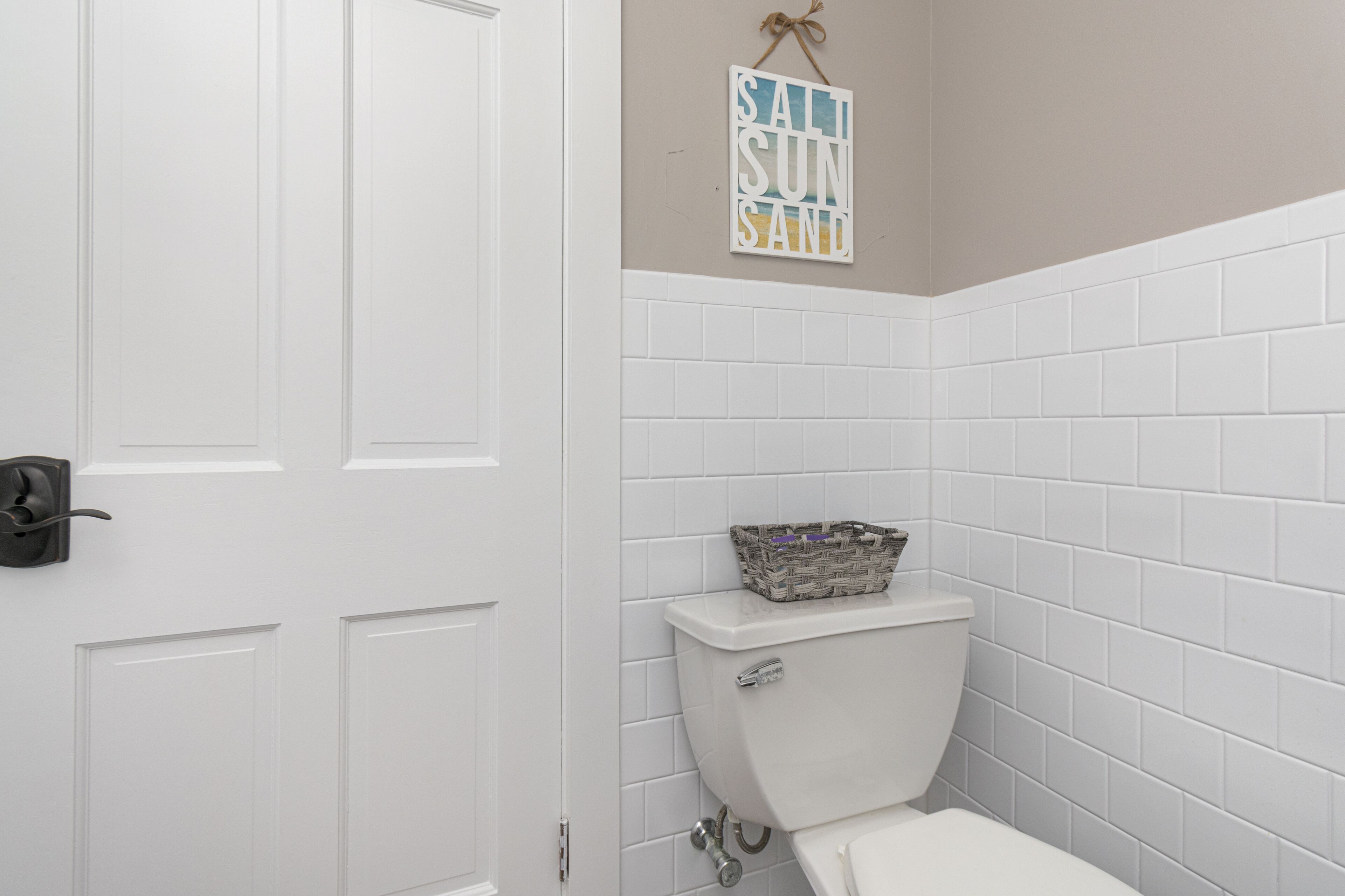 A bathroom with white tiled walls, a white toilet, and a basket on the toilet tank.