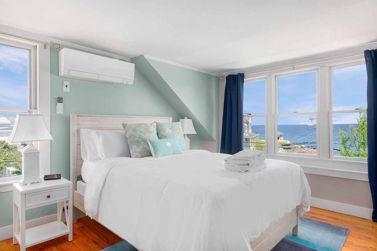 A bedroom with a bed, nightstand, lamp, and a view of the sea.