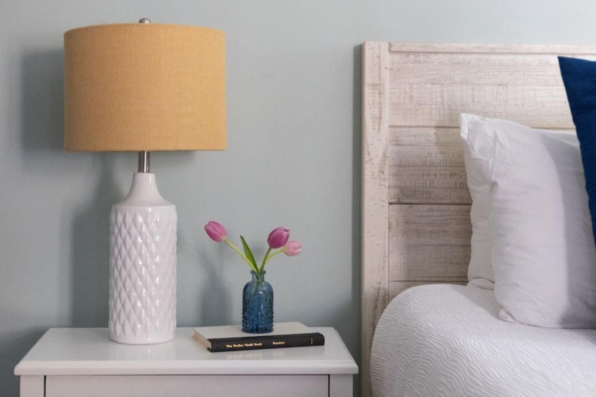 A neatly made bed with a blue pillow, a white lamp, and a vase with flowers on a bedside table.