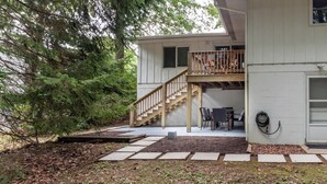 Una casa con una escalera de madera que lleva a una terraza, un patio con mesa y sillas, y un jardín con piedras para caminar.
