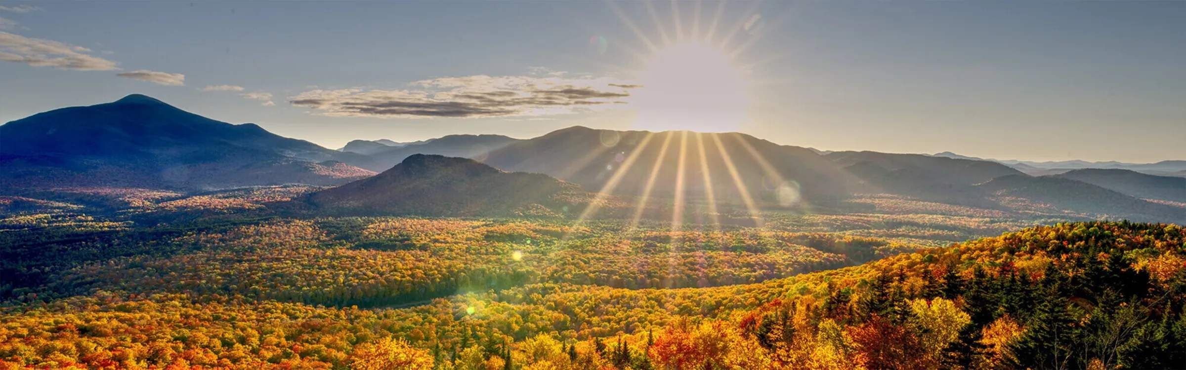 A mountainous landscape with a forested valley and a sun shining through the trees.