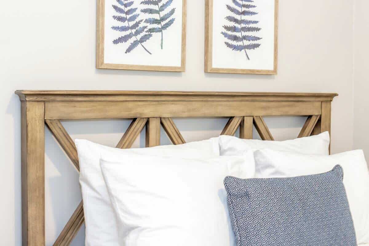 Een bed met een houten hoofdbord, witte en gemêleerde kussens, en twee ingelijste botanische prints aan de muur.