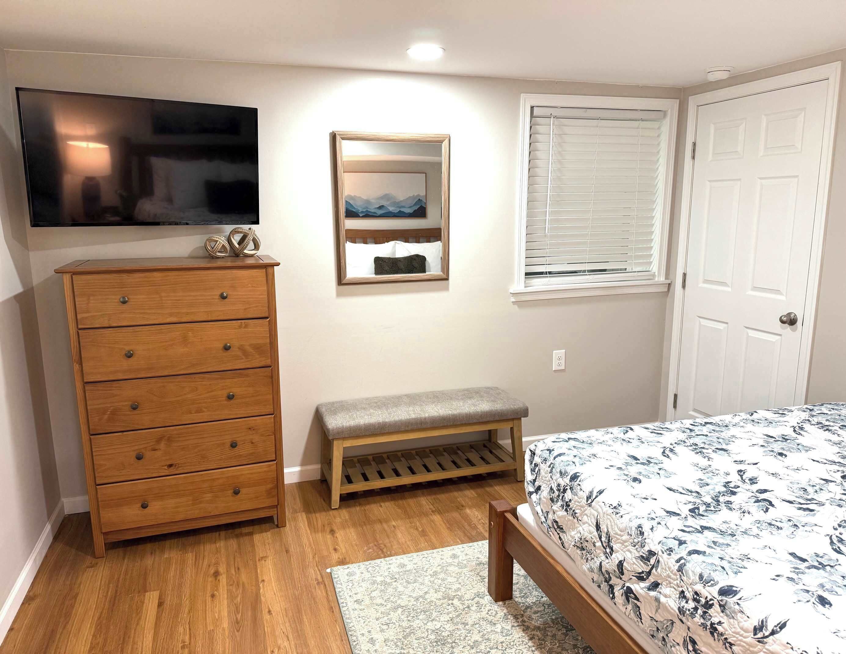 A bedroom with a wooden dresser, a bench, a bed with a floral bedspread, a mounted TV, a mirror, and a window with blinds.