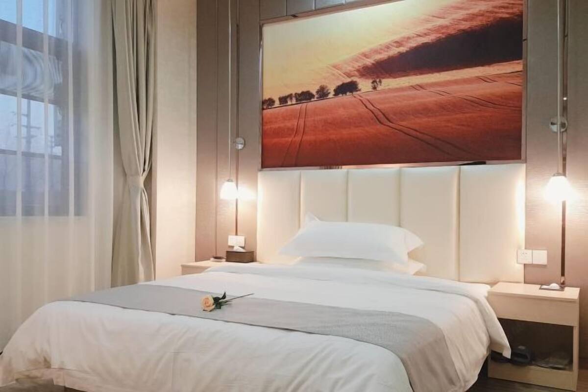 Een hotelkamer met een groot bed, twee bedlampen, een nachtkastje en een groot raam met dunne gordijnen.