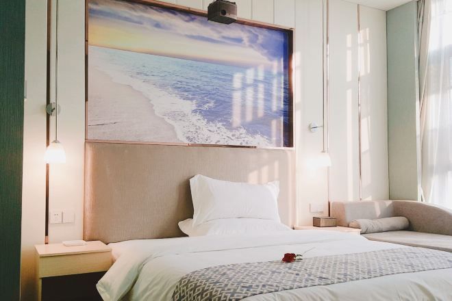 A hotel room with a bed, bedside tables, a nightstand, a wall-mounted lamp, and a large painting of a beach scene.
