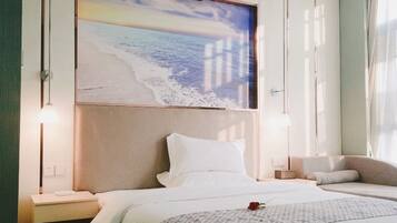 A hotel room with a bed, bedside tables, a nightstand, a wall-mounted lamp, and a large painting of a beach scene.