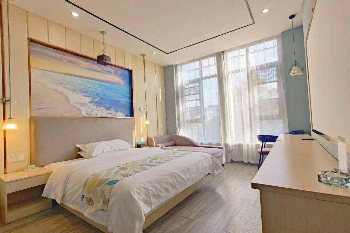 Een slaapkamer met een bed, nachtkastjes, een bureau, een groot raam en een schilderij aan de muur.