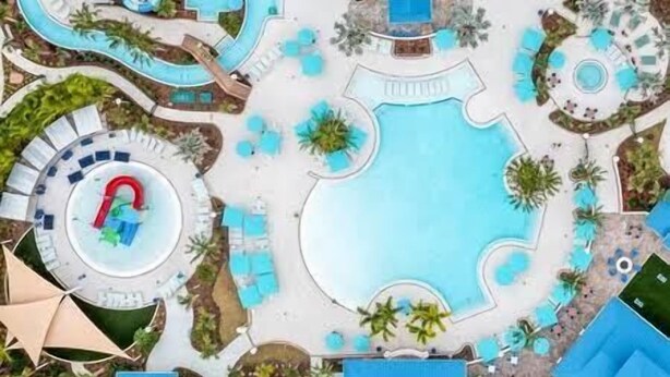 Vista aérea de un complejo turístico con una gran piscina central, varias piscinas más pequeñas y diversas fuentes acuáticas.