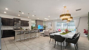 Una cocina moderna con armarios oscuros, una isla central con taburetes de bar, un comedor con mesa y sillas blancas, y una lámpara de araña grande.
