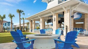 A covered outdoor seating area with blue Adirondack chairs, a fire pit, and a poolside bar.