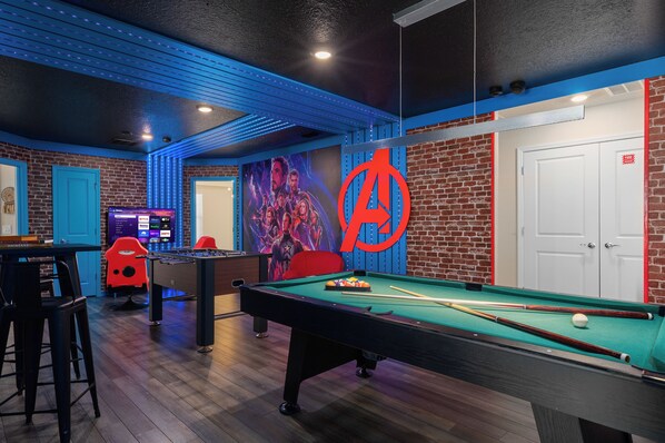 A pool table, a foosball table, and a large wall mural in a room with brick walls and blue lighting accents.