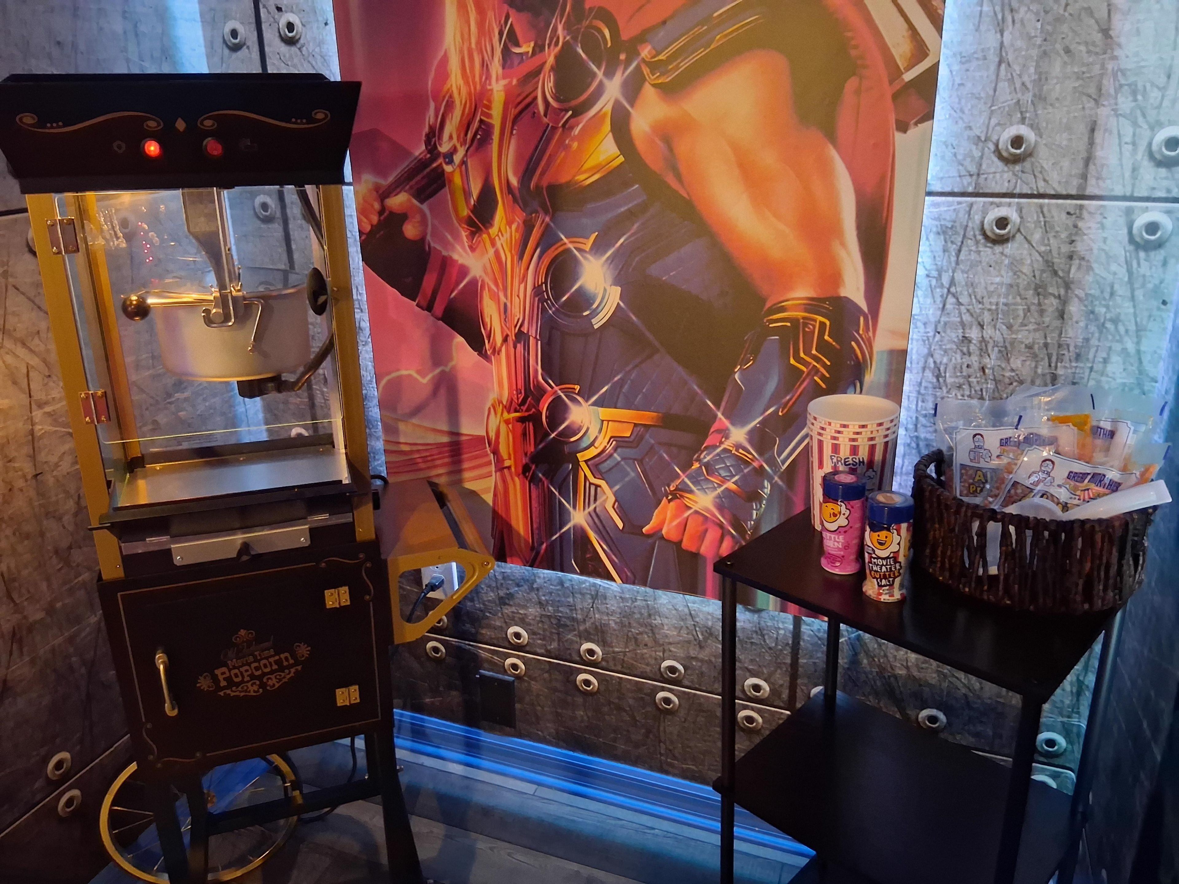 A popcorn machine with a branded sign, a wall with a vibrant mural, and a table with snack bags and a drink.