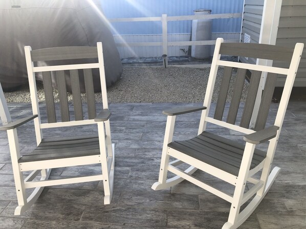 Dos mecedoras con estructura blanca y asientos de madera oscura, ubicadas en un patio de baldosas con zona de grava y una valla al fondo.