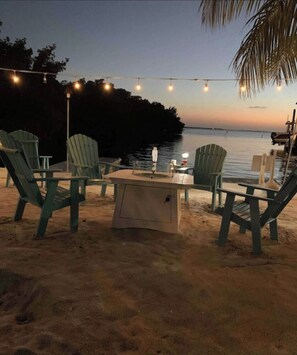 Un área de estar frente a la playa con sillas Adirondack y una mesa, luces colgantes y vista al agua al atardecer.