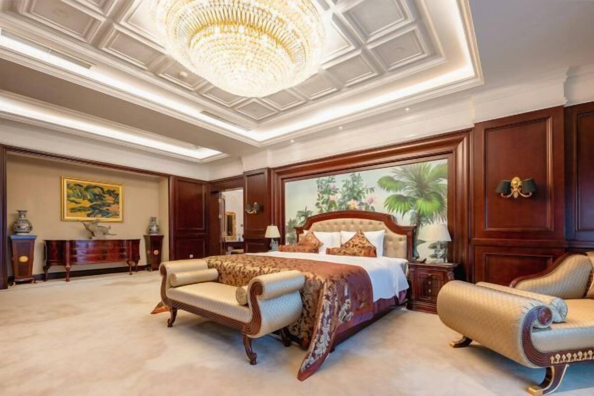 A spacious bedroom with a large bed, a chaise lounge, a chandelier, and a large window with a tropical view.