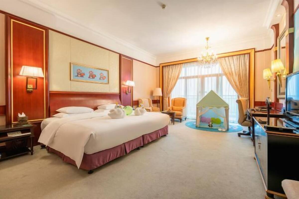 A hotel room with a large bed, two bedside lamps, a desk with a computer, a chair, a small play tent, and a window with curtains.
