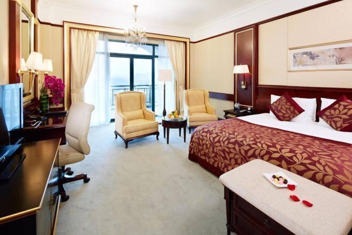 A hotel room with a bed, two armchairs, a desk, a TV, and a balcony with curtains.