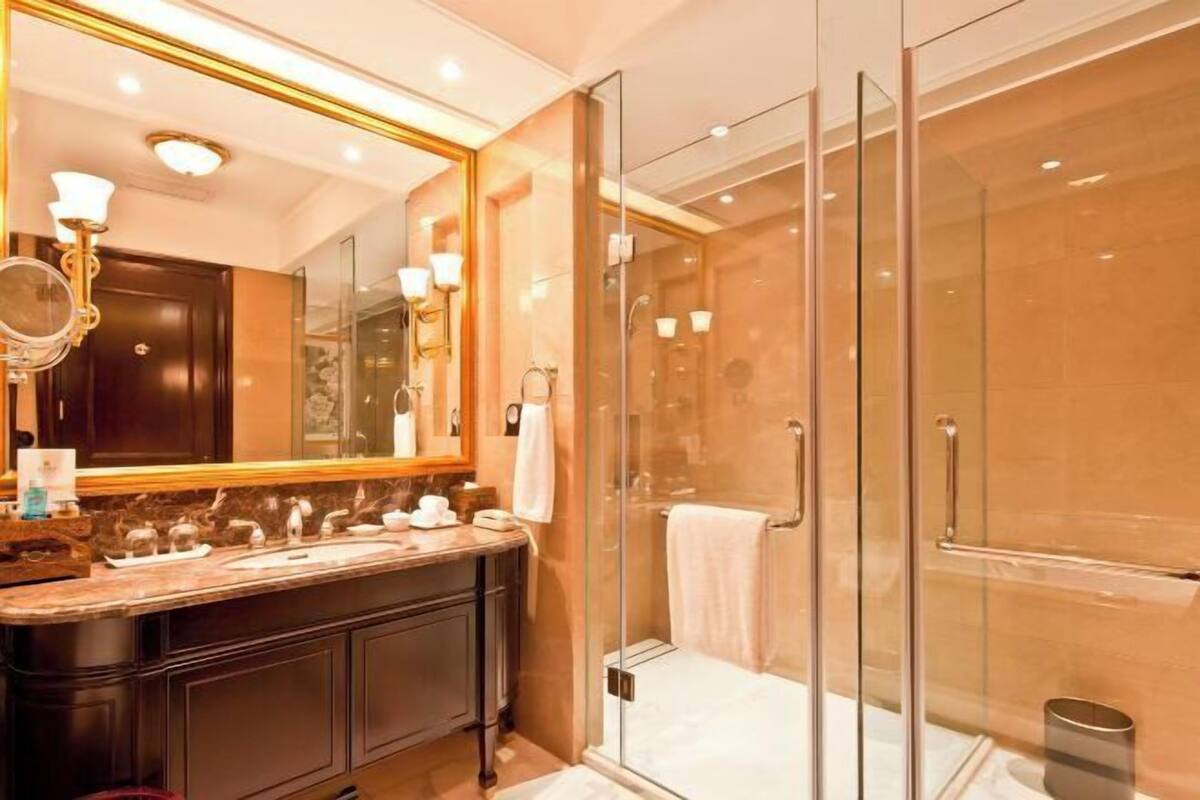 A modern bathroom with a glass-enclosed shower, a large mirror with a gold frame, and a marble countertop.
