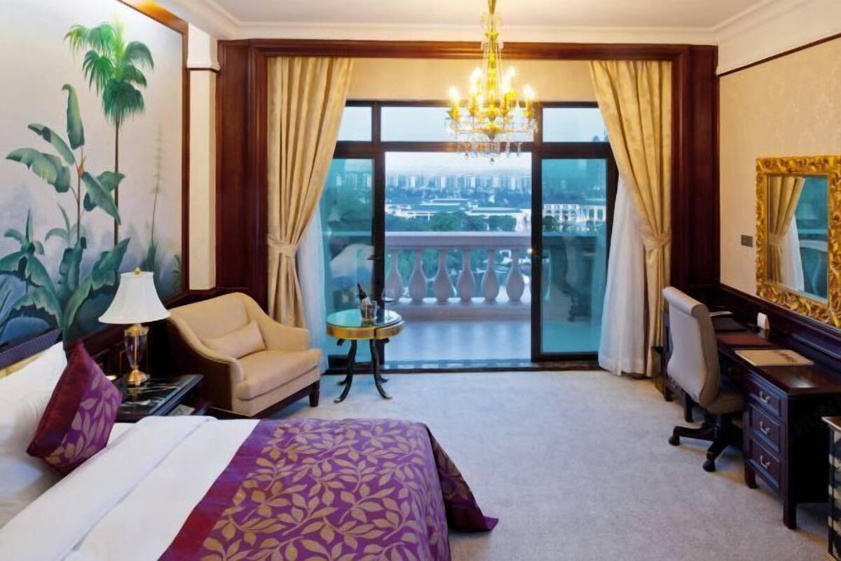 A hotel room with a large bed, a desk, a chair, a small table, a chandelier, and a balcony with a city view.