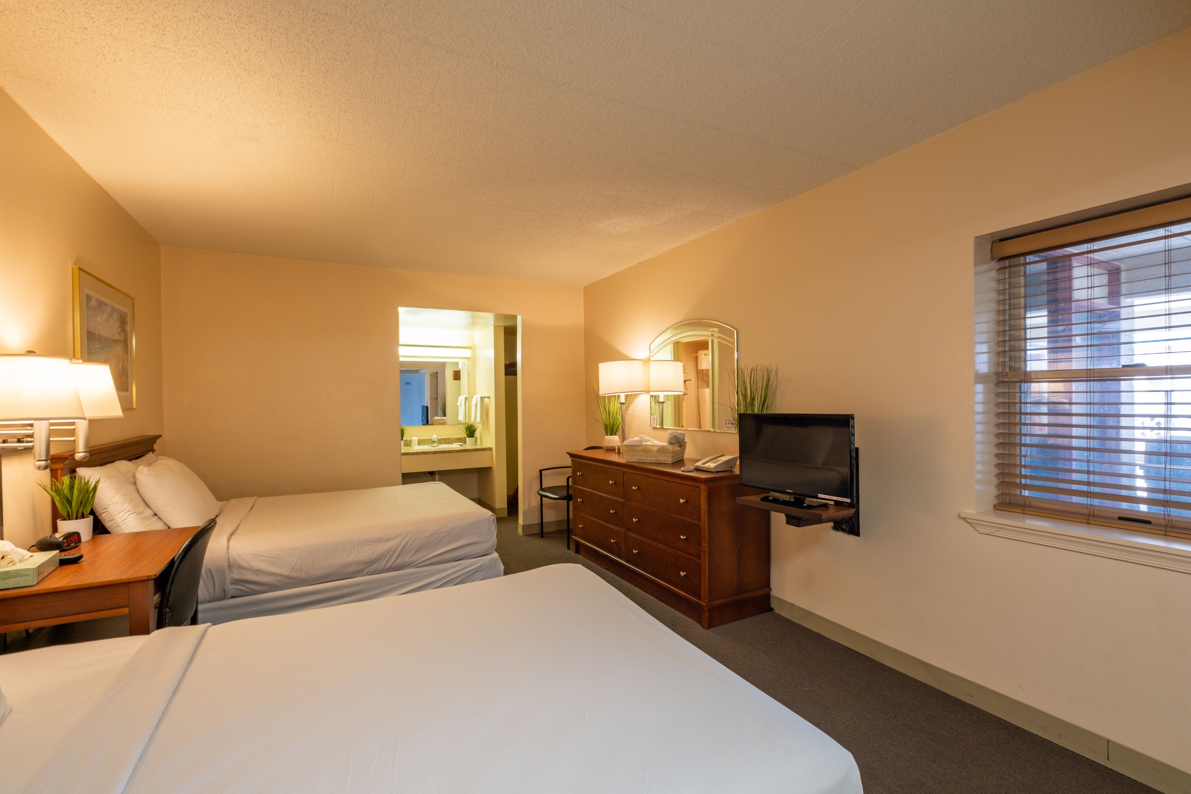 A hotel room with two beds, a dresser, a TV, a desk with a lamp, and a window with blinds.