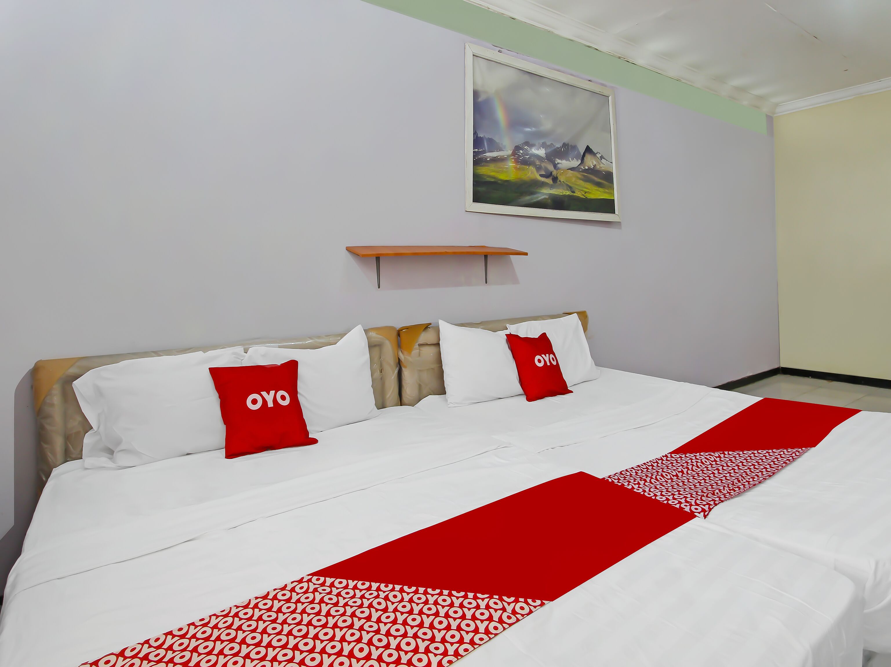 Quarto de hotel com cama, roupa de cama branca, almofadas vermelhas, corredor com padrão vermelho e branco, pintura de paisagem montanhosa e pequena prateleira de madeira.