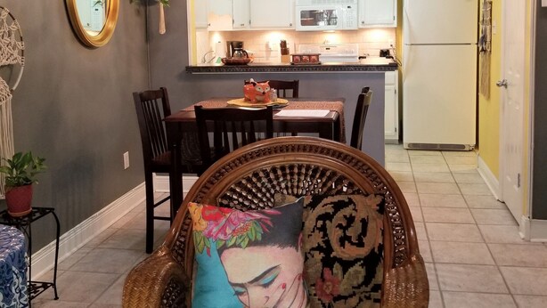 A dining area with a wicker chair, a table, and chairs. There is a kitchen in the background with white cabinets and a refrigerator.