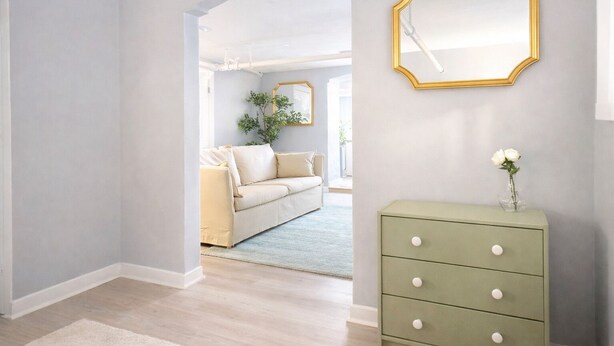 Ein Zimmer mit hellblauer Wand, einer grünen Kommode mit Schubladen, einer weißen Vase mit Blumen und einem großen Spiegel mit goldener Rahmung.