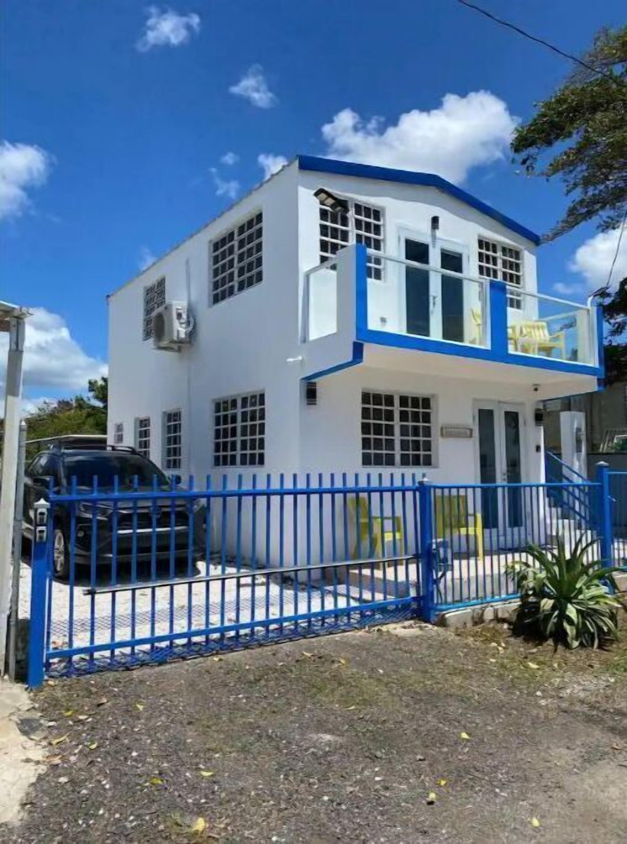 Un edificio de dos plantas con fachada azul y blanca, una entrada protegida por una reja y un coche estacionado.