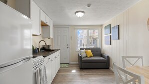 Cocina compacta con gabinetes blancos, estufa y lavamanos. Un sofá gris con una almohada amarilla, una mesa de comedor con sillas blancas y una ventana con vistas a edificios.