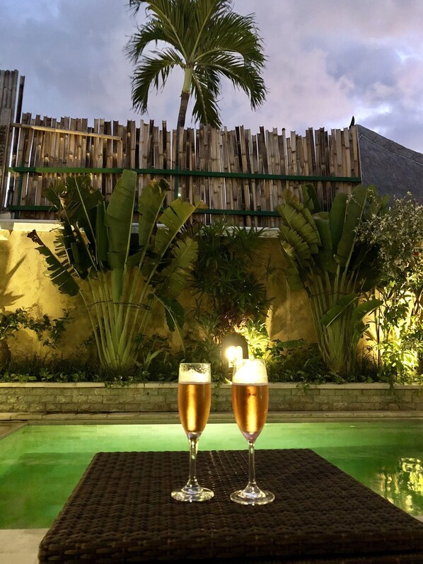 Deux verres de bière sur une table en osier, dans un cadre extérieur agrémenté d’une clôture en bois et de plantes tropicales en arrière-plan.
