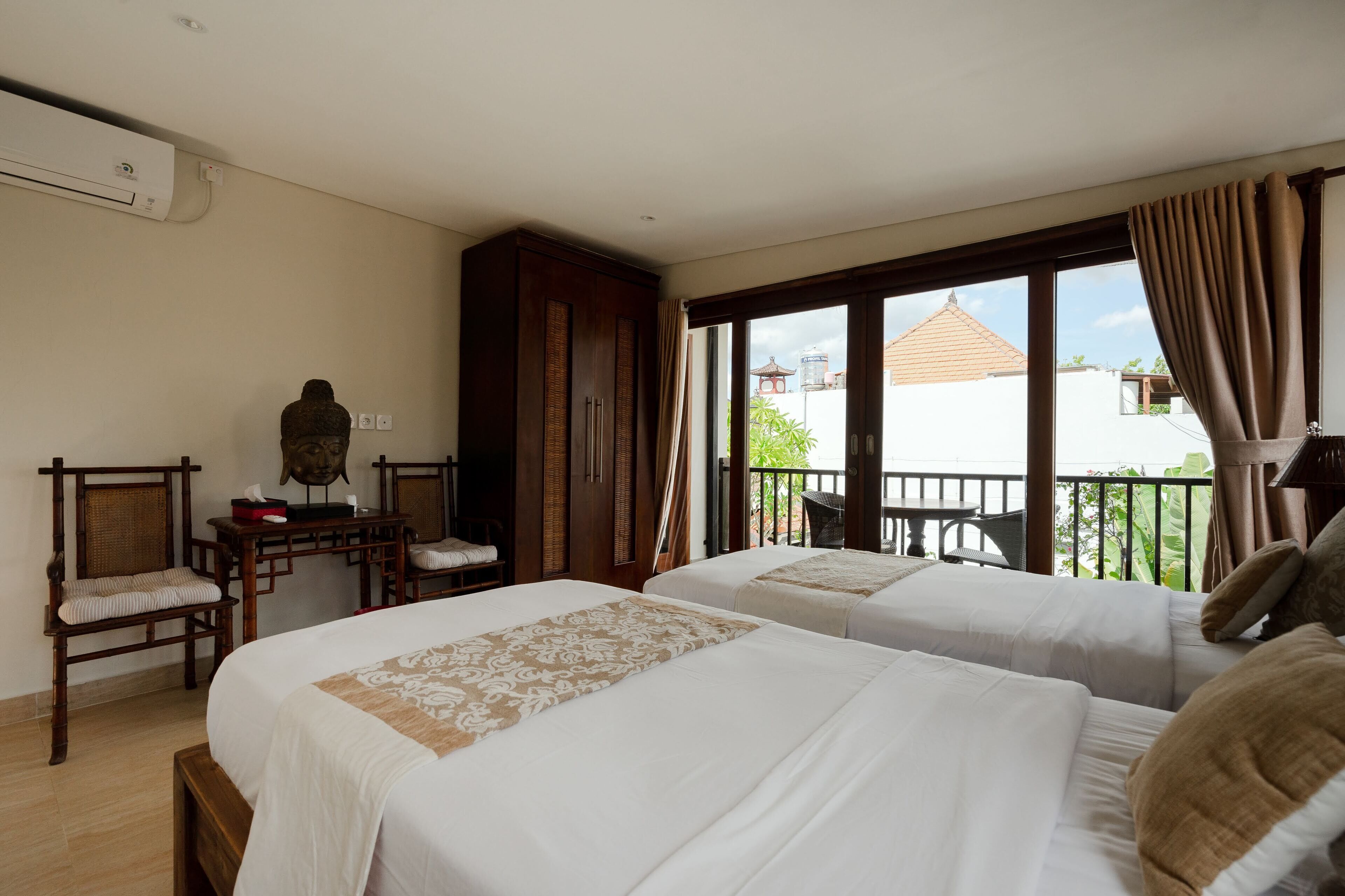 Habitación de hotel con dos camas, un escritorio de madera, una silla y un balcón con vistas a edificios.